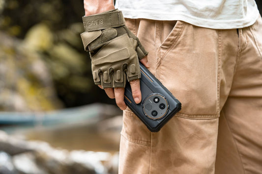 Gloved person carrying a black rugged Oukitel smartphone beside an outdoor stream.